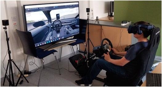 The study setting. The study participant is wearing the HTC Vive Pro Eye Virtual Reality headset, which includes speakers for audio output. The Logitech steering wheel and pedals are used for take-overs and manual driving. The microphone next to the steering wheel is used for speech input. The monitor is used by the study organizer to follow along.